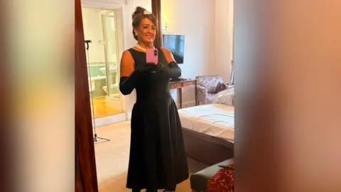 Lisa Hughes Woman wears a brunette updo wig and long black dress with black gloves and a pearl necklace.