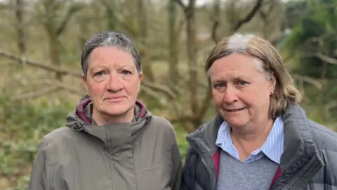 Two residents wearing coats standing in front of the stream with green foliage around them, one has short dark hair and the other shoulder length brown hair