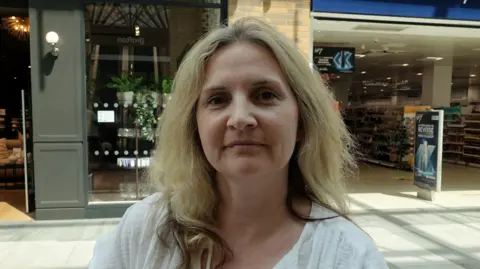 Head-and-shoulders photo of Sarah Lefever wearing a white top, with blonde hair, and shops in the background.