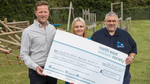 North East Autism Society Robert Poole, Corrina McEwan and Tony Wedlake of the Teesside Family Foundation, standing on a grass field, with a wooden playground behind them, with a cheque which reads for a value of £10,000.
