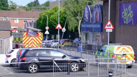 A red and yellow fire engine is parked outside the main entrance of Dundonald Ice Bowl. A green and yellow emergency vehicle is parked close by. A number of cars are parked in the car park around the building.