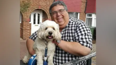 Councillor Richard Udall stands in a garden outside a red brick property. He wears glasses and a short-sleeved checked shirt. He is holding his white dog which looks like a poodle-cross.