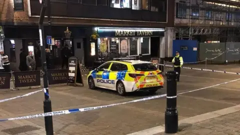 A yellow police car and police officer are standing in a cordoned-off area of Croydon, a pub called the Market Tavern is visible in the background