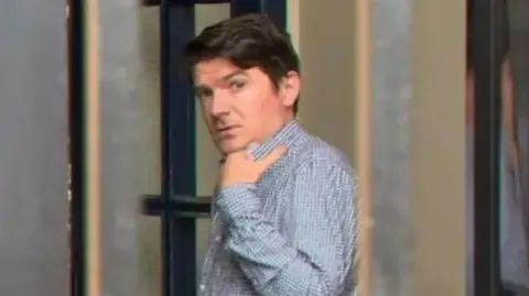 Michael McMonagle - a man with brown hair wearing a blue and white checked shirt looks at the camera as he walks towards a glass door