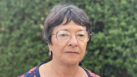Luke Deal/BBC Margaret Berrill looks at the camera while standing in front of a green bush. She has short dark hair with some grey in it. She wears round glasses and a navy top with a red floral pattern on it. 