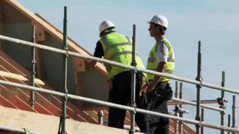 Getty Images Two workmen standing on scaffolding looking at the roof of a house