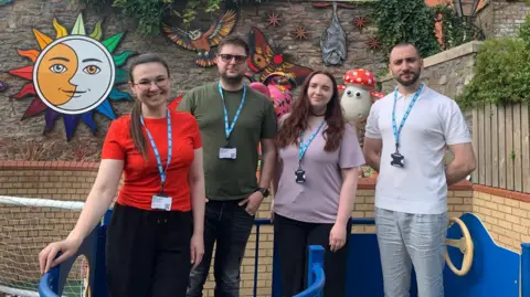 UHBW A group of four young Ukrainian medics, two men and two women, stand in a brick-lined play area in a line for the camera outside the Bristol Children's Hospital.