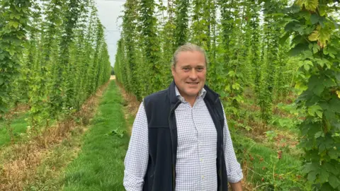 Emily Coady-Stemp/BBC An image of Matthew King, the estate manager at the Hogs Back Brewery in Tongham, in Surrey. Matthew is standing in a field and is wearing a checked shirt and gilet. His hair is tied back.
