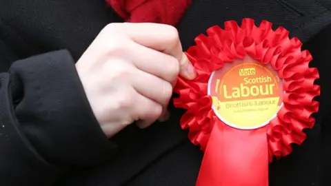 A person holds a red Labour rosette pinned to their black jacket 