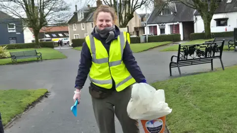 Renata Kaminsk, a smiling woman with brown hair and a hi-vis jacket, carrying a large orange bucket in one hand and a blue scoop in the other