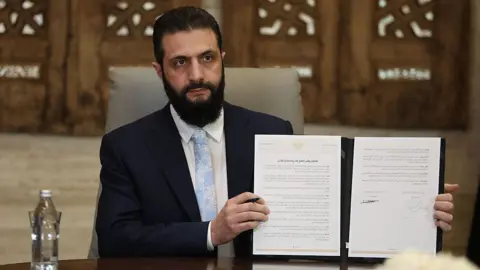 Syrian President Ahmed Sharaa in a navy suit, pale blue tie and white shirt holding up a physical copy of the agreement 