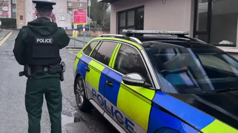 A police man with his back turned with his hand on his earpiece. He is wearing a full green PSNI outfit and has two arms in his back pockets. He is standing beside a yellow and blue police car. 