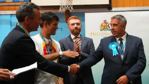 Getty Images Four people at an indoor election count stand in front of a “Hillingdon London” sign and a basketball hoop, as Steve Tuckwell in a suit wearing a blue rosette shakes hands with Ed Gemmell in a dark suit, while two others look on and clap.