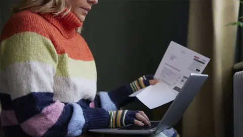 Getty Images A woman sits in her home wearing a stripey jumper and gloves whilst reading a bill with her laptop on her lap