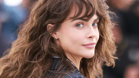 Cara Delevingne wears a navy pinstripe jacket. She is looking over her shoulder and has brown wavy hair.