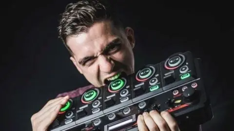Nue A man is holding loopstation equipment in his hands, which he jokingly bites with his teeth. The equipment is black and grey with lots of knobs and buttons that are partly lit up in green and red.