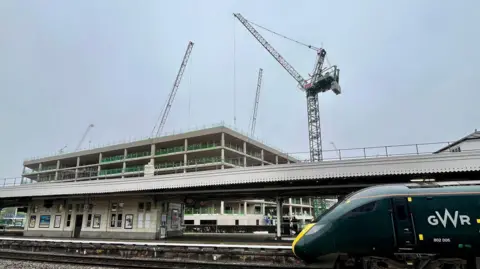 Cranes are visible on a large building site next to Temple Meads Station Bristol, with two stories of a new building visible. In the foreground is a Great Western Railway train with dark green livery waiting at a platform.