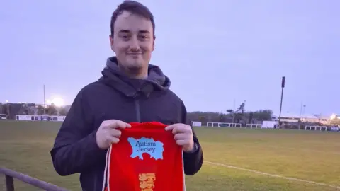 A picture of a man holding a red bag which reads 'Autism Jersey' on it. It is red and light blue in colour.