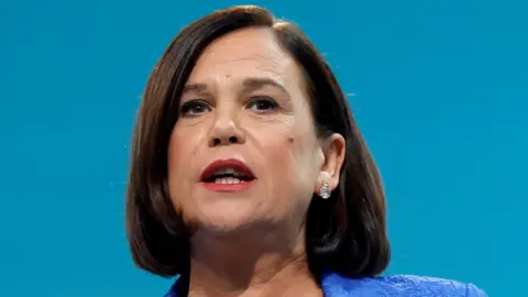 Reuters Mary Lou McDonald - a woman with dark, short hair is speaking with her mouth open. She is wearing a blue suit jacket and is standing against a blue background.