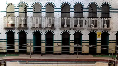 Getty Images The side of a swimming pool with a white and beige tiled wall. There are eight green tiled arched doorways, which have a semicircle of black and white tiling above each one. On the floor above are the same style of arched entrances, but they are protected by white metal balcony-style railings.