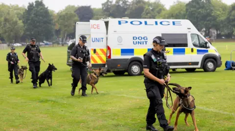 West Midlands PCC Four police officers in black uniform walk across a green field, each of them holding the lead of a police dog who walks alongside them. A white police dog unit van is behind them.
