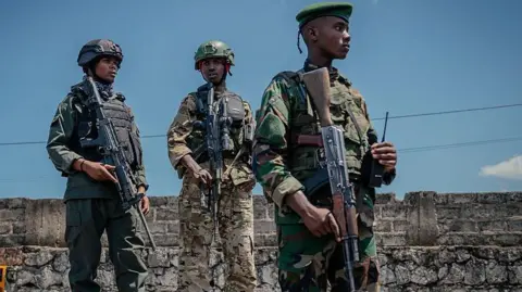 Three men in military fatigues and holding rifles stand in front of a breeze block wall.