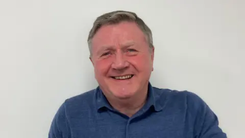 Dennis Jones smiling at the camera against a white wall, wearing a blue polo shirt.