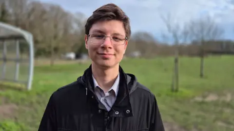 Stuart Woodward/BBC Kiril Vorobiov wears a black jacket, open at the neck, with a light blue shirt underneath. He wears thin grey-rimmed spectacles and has brown hair in a side parting. He is standing on an area of grass with trees and blue sky behind.