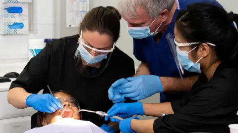 University of Lancashire Two female dentists with dark hair tied back look into the mouth of a patient in a chair, while an older male dentist in navy scrubs watches. They are all wearing masks and blue latex gloves.
