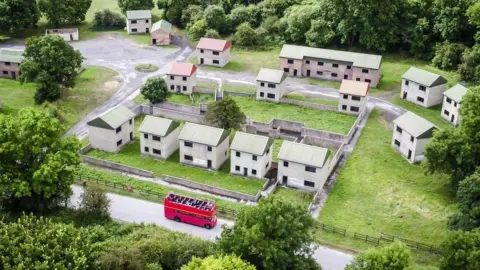 An aerial picture of what the village looks like today, with a small cluster of empty brick houses with tin roofs and no windows or doors. It is a stark and bare street and all the houses look identical. At the bottom there is a red double-decker tour bus driving along the road.