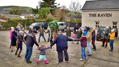 Manx Scenes Landscape shot of people celebrating Hunt the Wren. People have joined hands and formed a circle around a decorative pole on which the Wren sits.