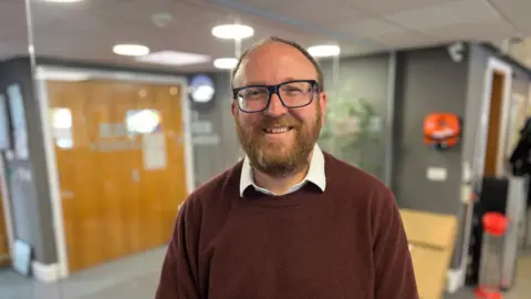 Deputy Garry Collins - A man with gingery brown hair wearing blue glasses looking at the camera. He has a thick brown beard.