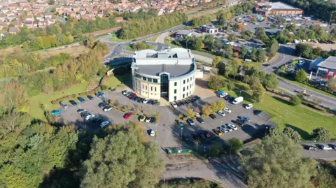 An aerial, drone-style photograph of a modern circular building surrounded by parking areas, greenery, and a mix of urban and suburban elements. The environment combines developed infrastructure with large patches of woodland and open green space. At the bottom of the image, dense clusters of mixed-height trees and shrubs fill the foreground. A road runs through or near the lower part of the frame, partly obscured by the trees.