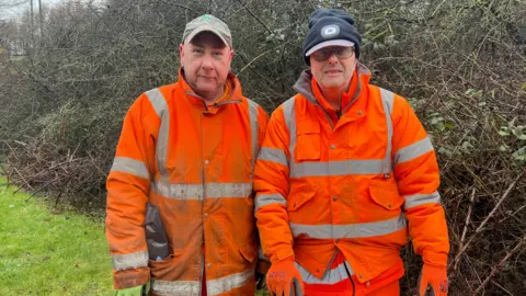Two men in orange high visibility gear stood in front of hedge 