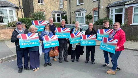 Reform UK An image posted by Reform UK which appears to have been modified to change the background behind a group of campaigners holding Reform UK signs. Behind them are houses with what appears to be part of a car behind the group and on the pavement. They are each smiling and holding up pale blue signs with a section of union jack flag on them, bearing the words "I'm voting Reform UK".