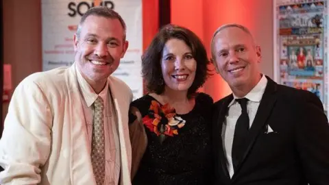 Meryl Praill, a dark-haired woman wearing a black dress and a red and orange corsage, Will Young, a grey-haired man wearing a stripy shirt and a while blazer, and Rob Rinder, and blonde man wearing a black tuxedo. All three are smiling at the camera and they're in a room with a red wall behind them.