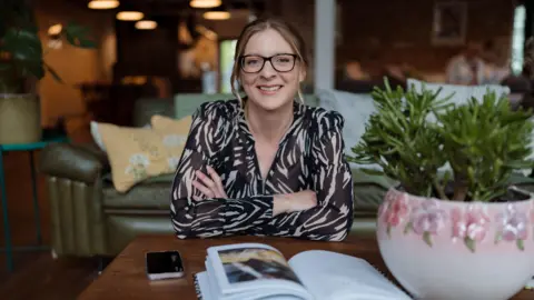 Trish Peters Mandy Mazliah is sitting at a desk smiling at the camera. She is wearing glasses and has her hair in a ponytail. She has her arms crossed and is wearing a zebra print blouse.