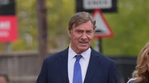 Lord Charles Brocket is pictured from the chest up, wearing a dark navy suit jacket, a white dress shirt, and a blue patterned tie. He is outdoors with a blurred background that includes green foliage and a red and white triangular road sign. He is looking slightly to his left with a neutral expression.