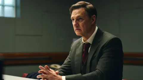 ITV Morrissey sat at a table in a police interview room, wearing a dark suit, white shirt and burgundy tie. He has short dark hair and has a worried expression. 