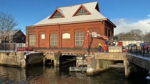BBC/Ian Forster Workers in high-visibility clothing stand at the edge of where a bridge once was across the River Wensum in Norwich, having removed it