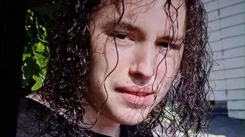 Family photo Vlad Nikolin-Caisley has curly, dark, hair down to his neck in a wet-look style. He is half-smiling and looks at a point a little to the left of the camera.