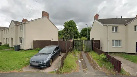 A pathway leads between two houses to a gate, partially open and which appears to lead to an area with greenery. A car is parked on the driveway of a nearby house.