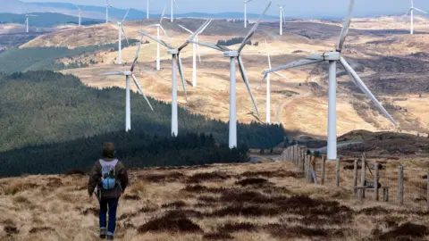 Wind turbines at Cefn Croes Wind Farm on 16th March 2025 in Cwmystwyth, Wales, United Kingdom. Cefn Croes is a wind farm in Ceredigion, located in the Cambrian Mountains.  A person can be seen walking in the foreground, wearing a dark brown coat and purple backpack.
