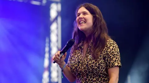 Rosie Jones in a leopard print jumpsuit looking off camera smiling and holding a microphone. She has brown hair and is standing on a stage.