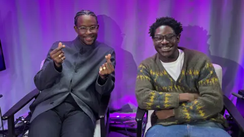 Two men sit on chairs in a studio with a purple curtain in the background. One man is dressed in a dark grey shirt, and smiles as he appears to click his fingers. The other man, who wears an olive cardigan with flashes of blue and yellow pattern, smiles widely with his arms folded in a relaxed posture.