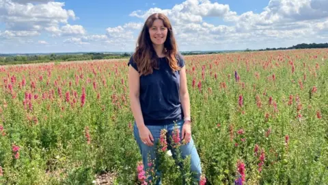 Ashley stood waist-deep in a field of dark pink larkspur flowers. She's wearing blue jeans and a dark blue tshirt. The sky is blue with fluffy clouds, and the sun is very bright. 