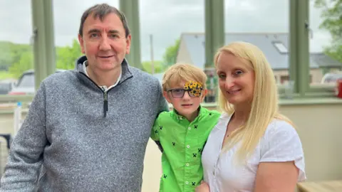 Vanessa Clarke / BBC Harry stands smiling in a green shirt wearing glasses and his patch which is orange with stars and rockets. His mum Ellen is on the right of the picture smiling with long blonde hair and his Dad Robert is on his right in a grey jumper.