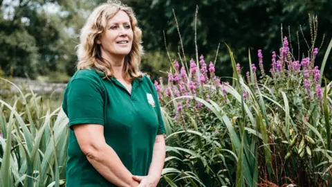 Future Roots Future Roots founder, Julie Plumley she is standing in front of flowers wearing a green top and has blond hair.
