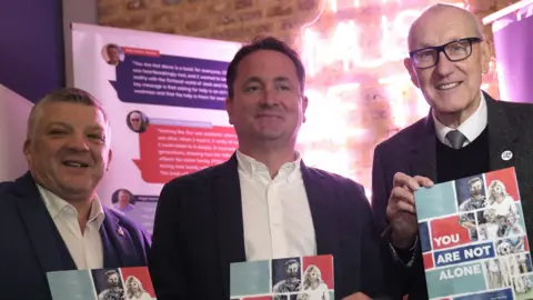 Nigel Seaman, Rob Parker and Terry Butcher stand together on a stage and smile at the camera. They each hold a copy of a book called You Are Not Alone and are wearing suit jackets with white shirts. 