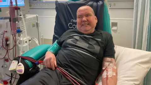 Anthony Devine is wearing a black t-shirt and a blood pressure cuff on his right arm, while the tubes needed to clean his blood through dialysis are draped across him and taped into he left arm.